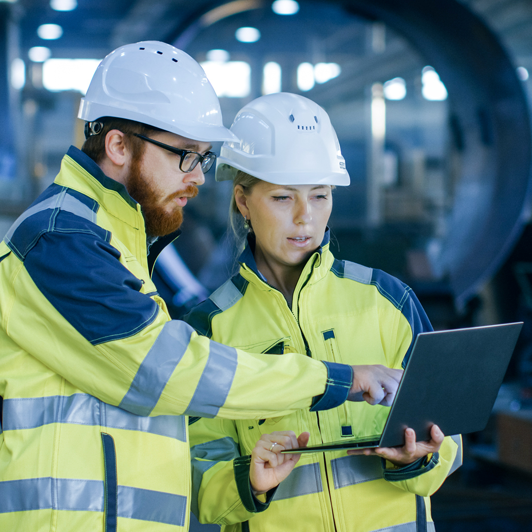 Bilde av mann og kvinne med hjelm på en fabrikk som ser på en PC